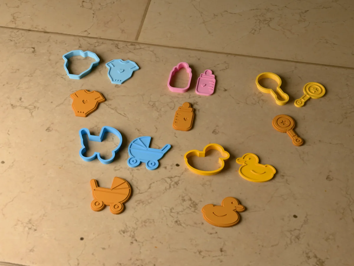 3D render of five 3D-printed baby shower cookie cutters with their stamps and a golden stamped cookie in front of each: onesie, baby bottle, rattle, pram and rubber duck