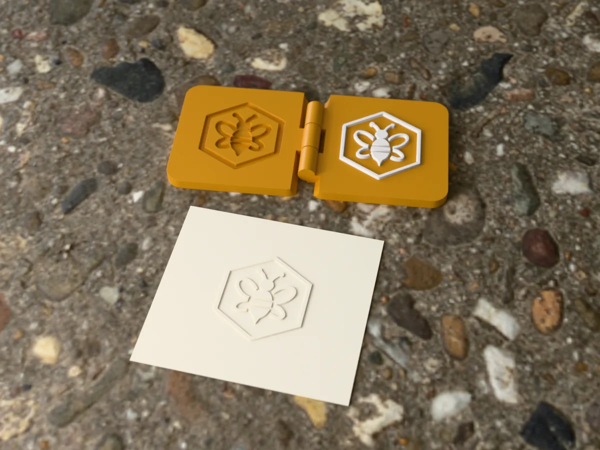 3D render of the printed bee embosser, open flat on its hinge, with the raised die and the matching cavity, next to a card embossed with its mark