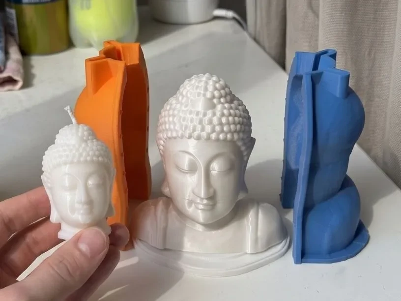 White wax Buddha head candle held in a hand, next to the white printed Buddha bust master and the orange and blue halves of the printed housing