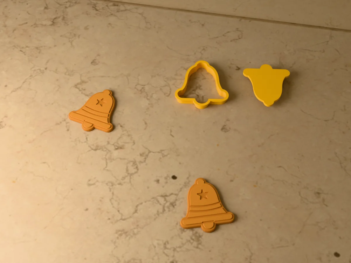3D render of a yellow 3D-printed Christmas bell cookie cutter and stamp next to two golden baked cookies with the bands, rim and a star pressed in