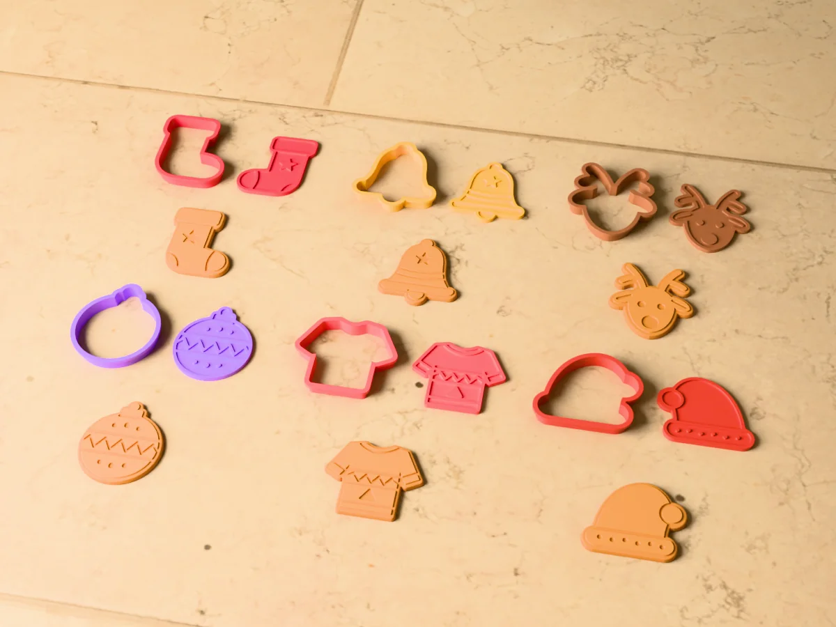 3D render of six 3D-printed Christmas cookie cutters with their stamps and a golden stamped cookie in front of each: Christmas stocking, Christmas bell, reindeer, Christmas ornament, Christmas sweater and Santa hat