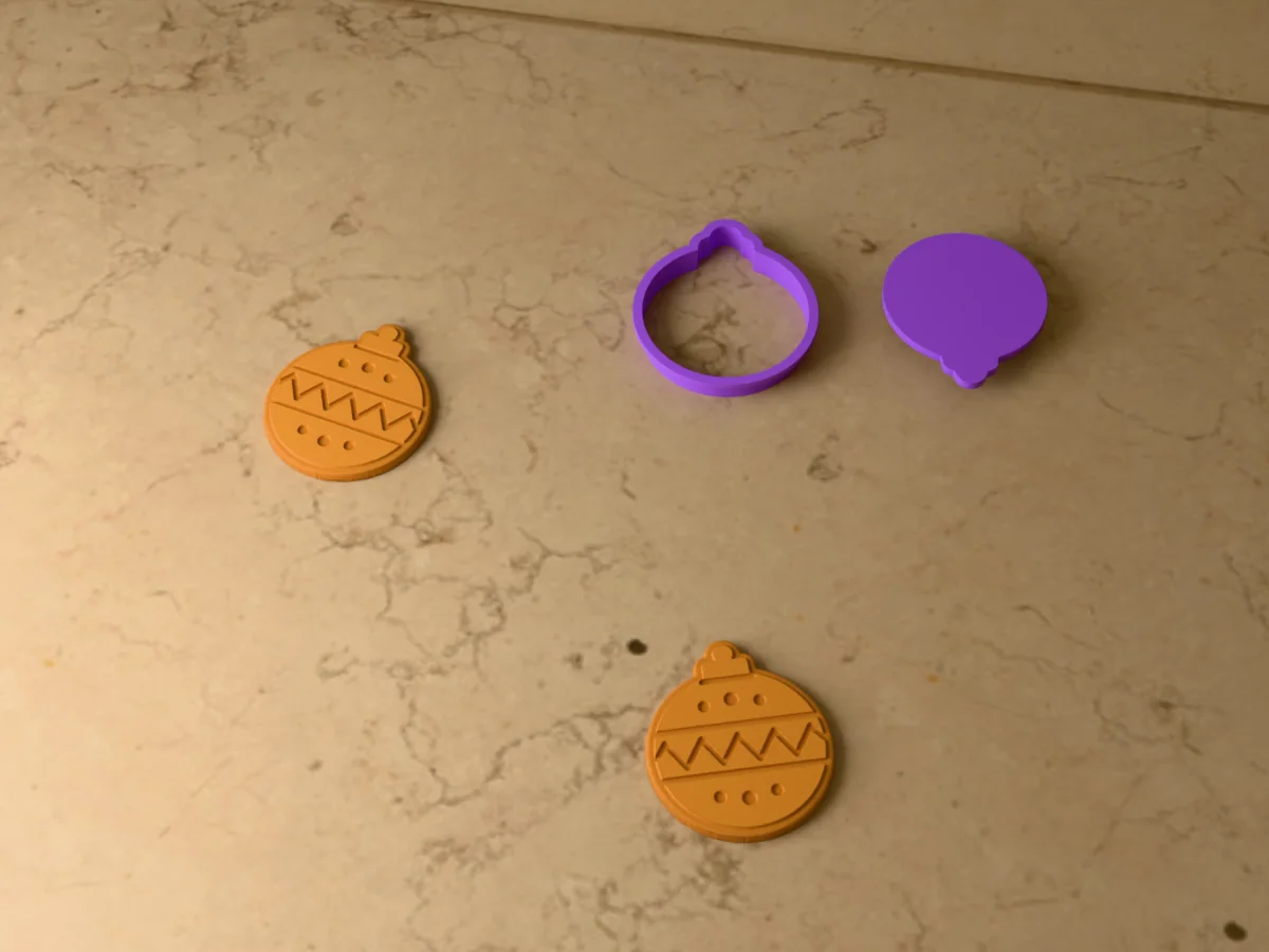 3D render of a purple 3D-printed Christmas ornament cookie cutter and stamp next to two golden baked cookies with bands, a zigzag and dots pressed in