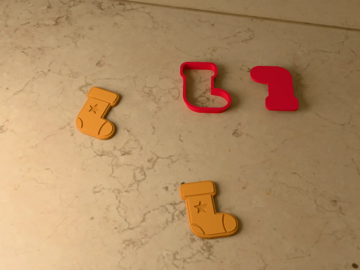 3D render of a red 3D-printed Christmas stocking cookie cutter and stamp next to two golden baked cookies with the cuff, heel, toe and a star pressed in