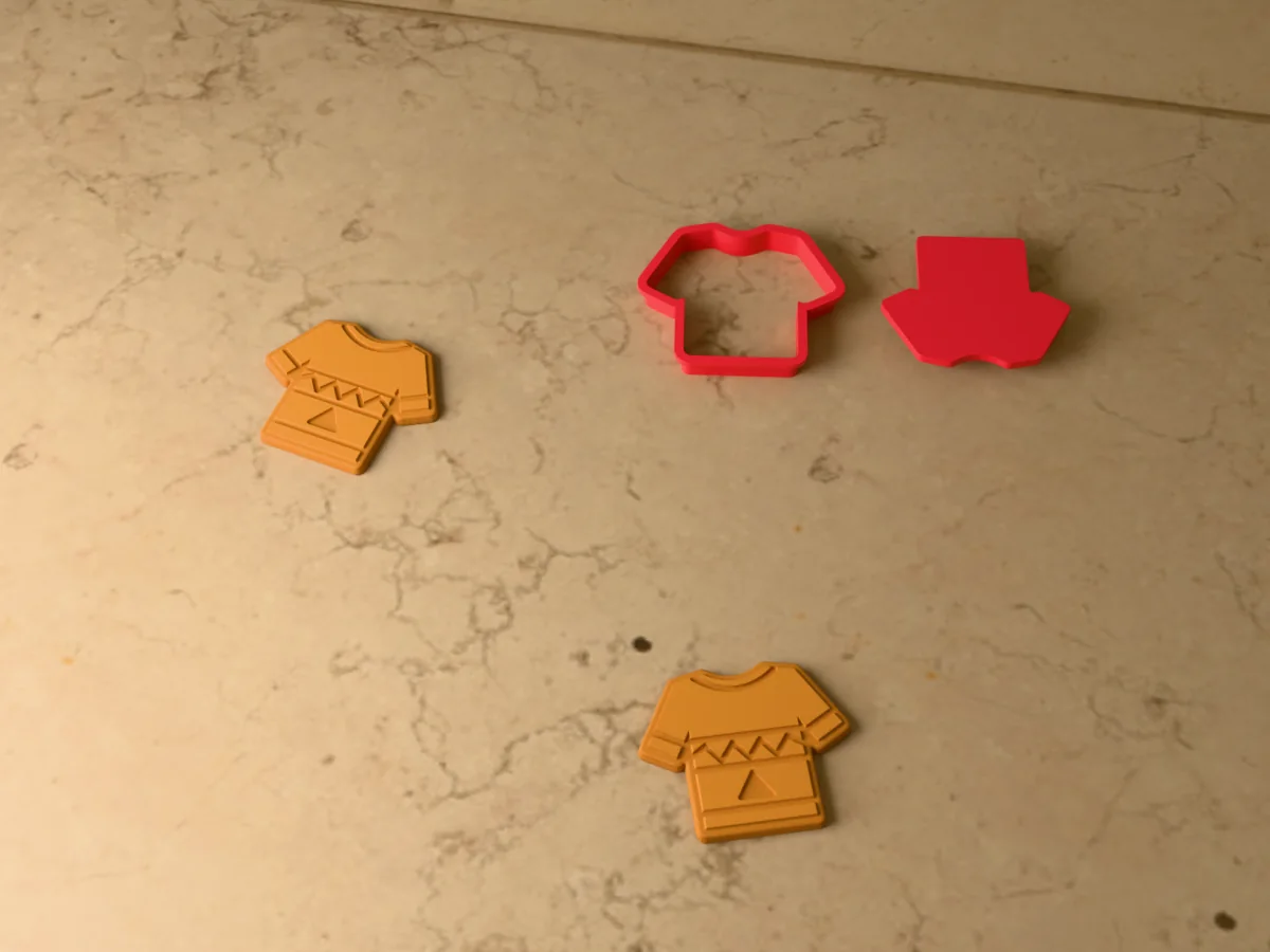 3D render of a red 3D-printed Christmas sweater cookie cutter and stamp next to two golden baked cookies with the ribbing, a zigzag band and a small tree pressed in