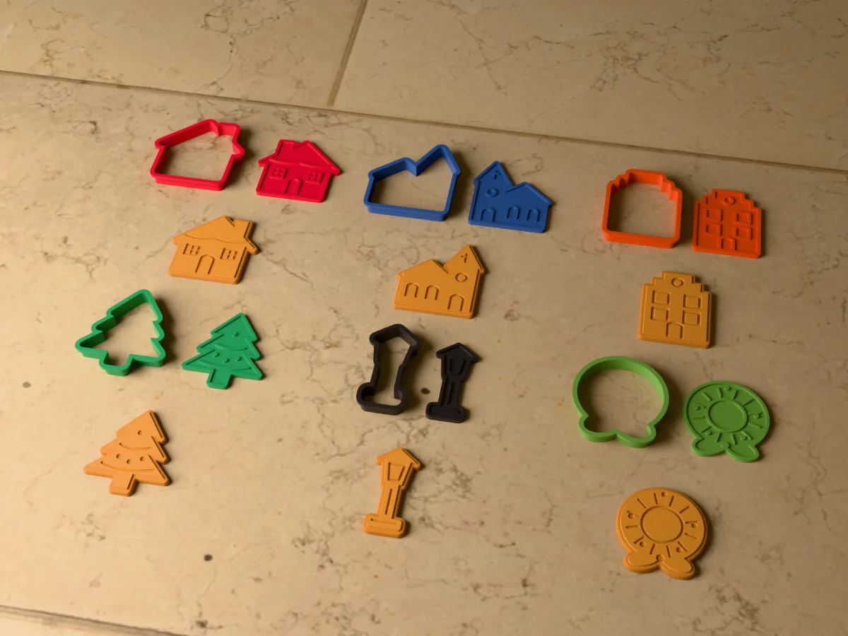 3D render of six 3D-printed Christmas village cookie cutters with their stamps and a golden stamped cookie in front of each: cottage, church, townhouse, fir tree, street lamp and wreath