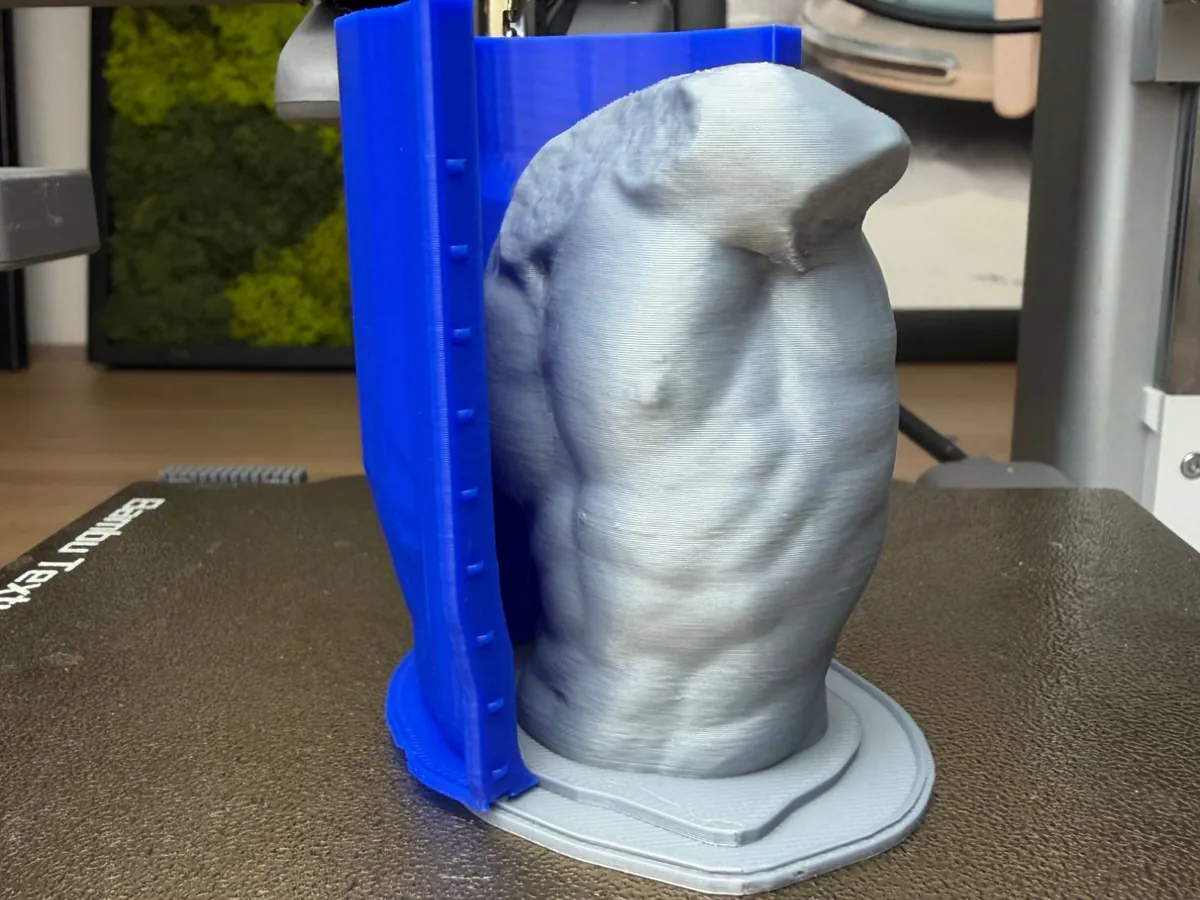 Printed grey classical torso fused on its base, with one blue hugging shell half standing behind it on a printer bed