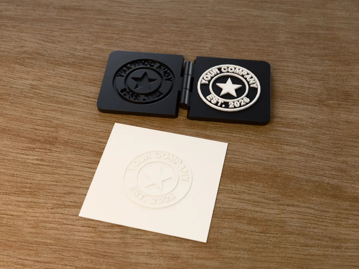 3D render of the printed company seal embosser, open flat on its hinge, with the raised die and the matching cavity, next to a card embossed with its mark
