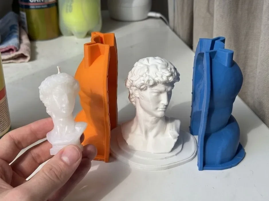 White wax David bust candle held in a hand, next to the white printed David bust master and the orange and blue halves of the printed housing