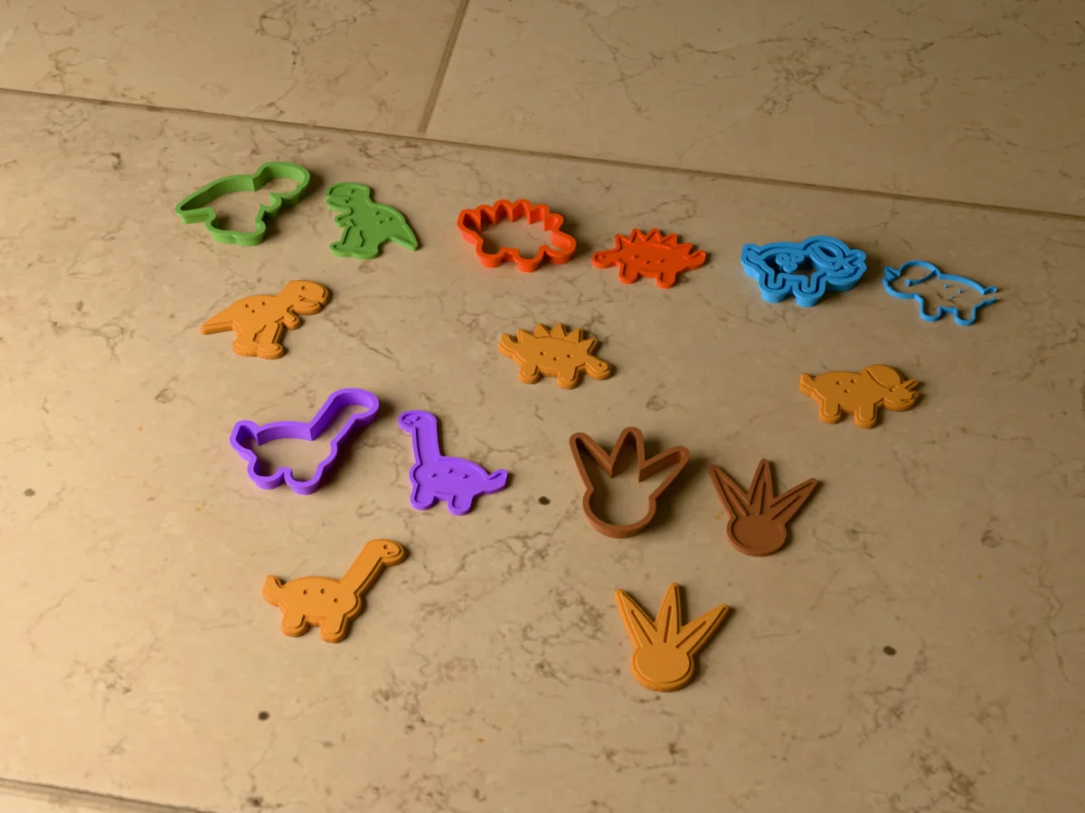 3D render of five 3D-printed dinosaur cookie cutters with their stamps and a golden stamped cookie in front of each: T-rex, stegosaurus, triceratops, long neck and dinosaur footprint
