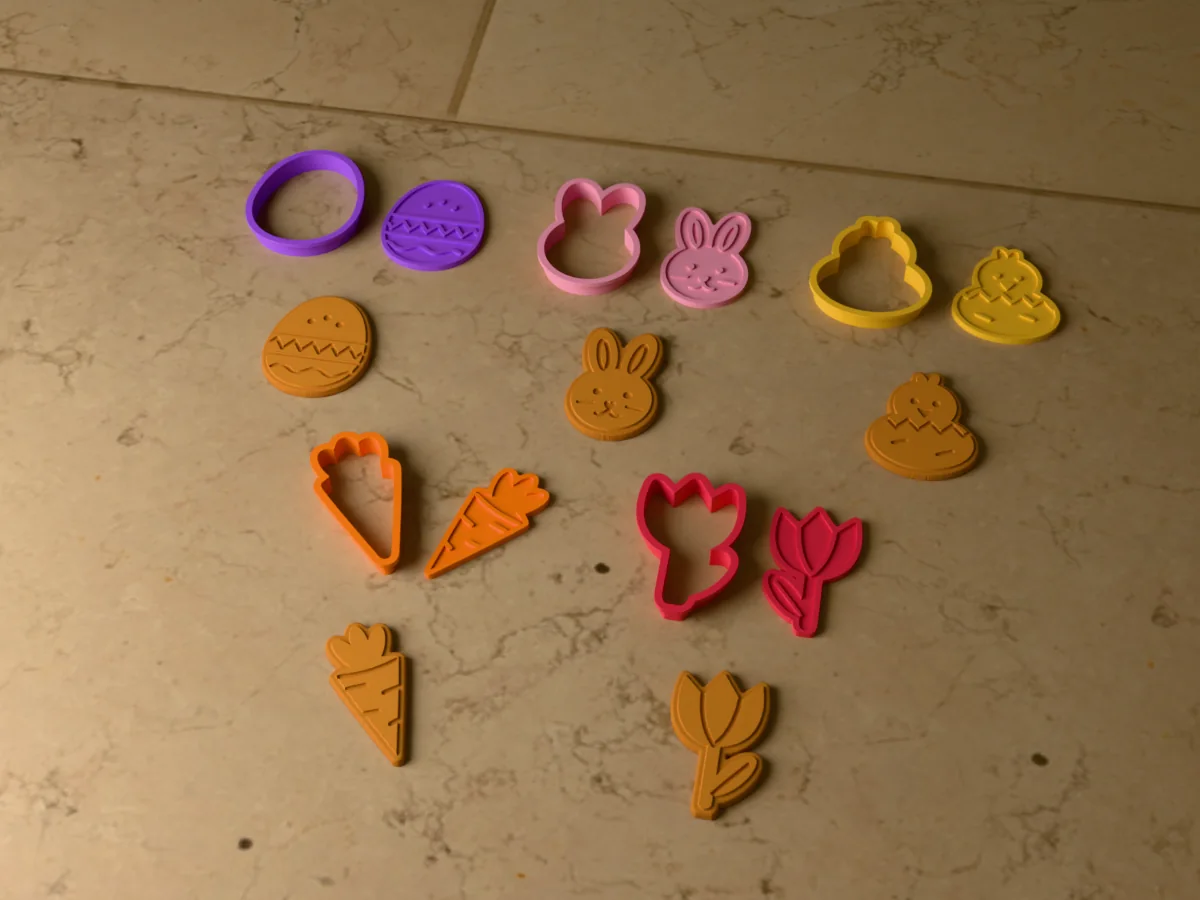 3D render of five 3D-printed Easter cookie cutters with their stamps and a golden stamped cookie in front of each: Easter egg, bunny, hatching chick, carrot and tulip