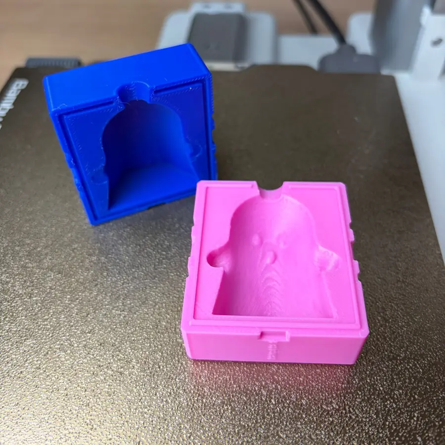 Both ghost mold halves on the build plate, the pink half lying open to show the face, arms and pour funnel pressed into the cavity