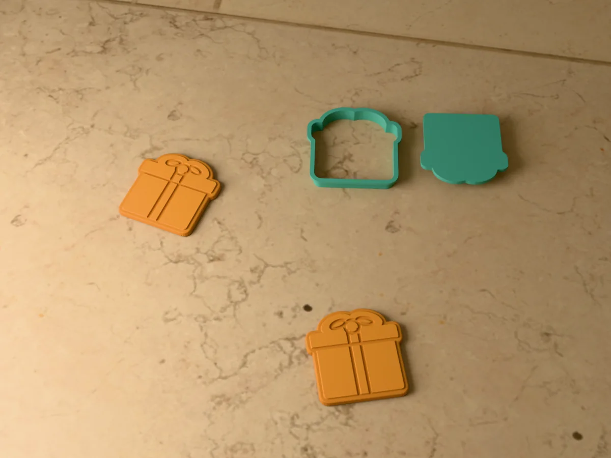 3D render of a teal green 3D-printed gift box cookie cutter and stamp next to two golden baked cookies with the lid, ribbon and bow pressed in