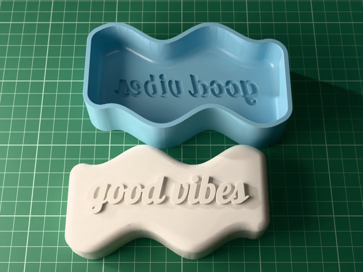 3D render from above of a white wavy slab candle lying face up with "good vibes" in raised script, the silicone mold it is cast in behind it