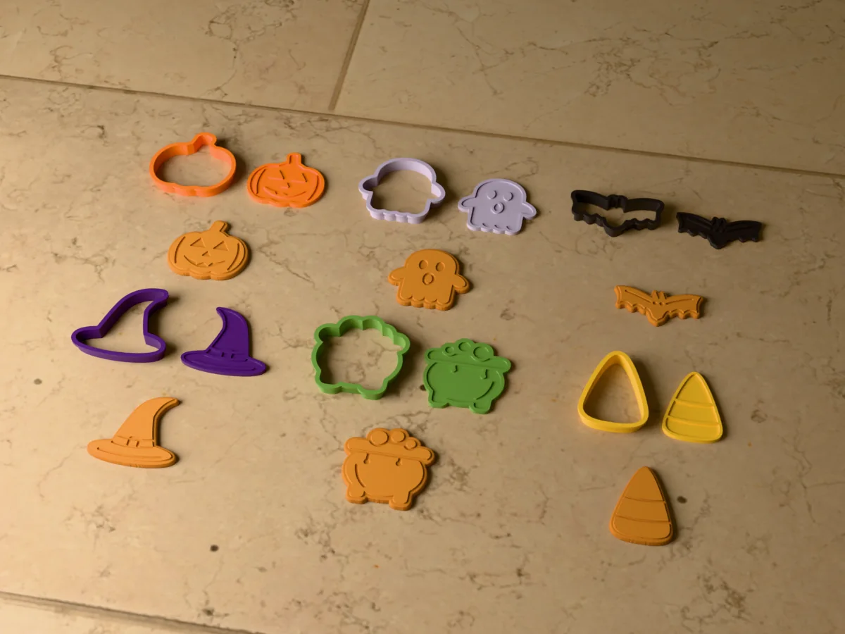 3D render of six 3D-printed Halloween cookie cutters with their stamps and a golden stamped cookie in front of each: jack-o'-lantern, ghost, bat, witch hat, cauldron and candy corn