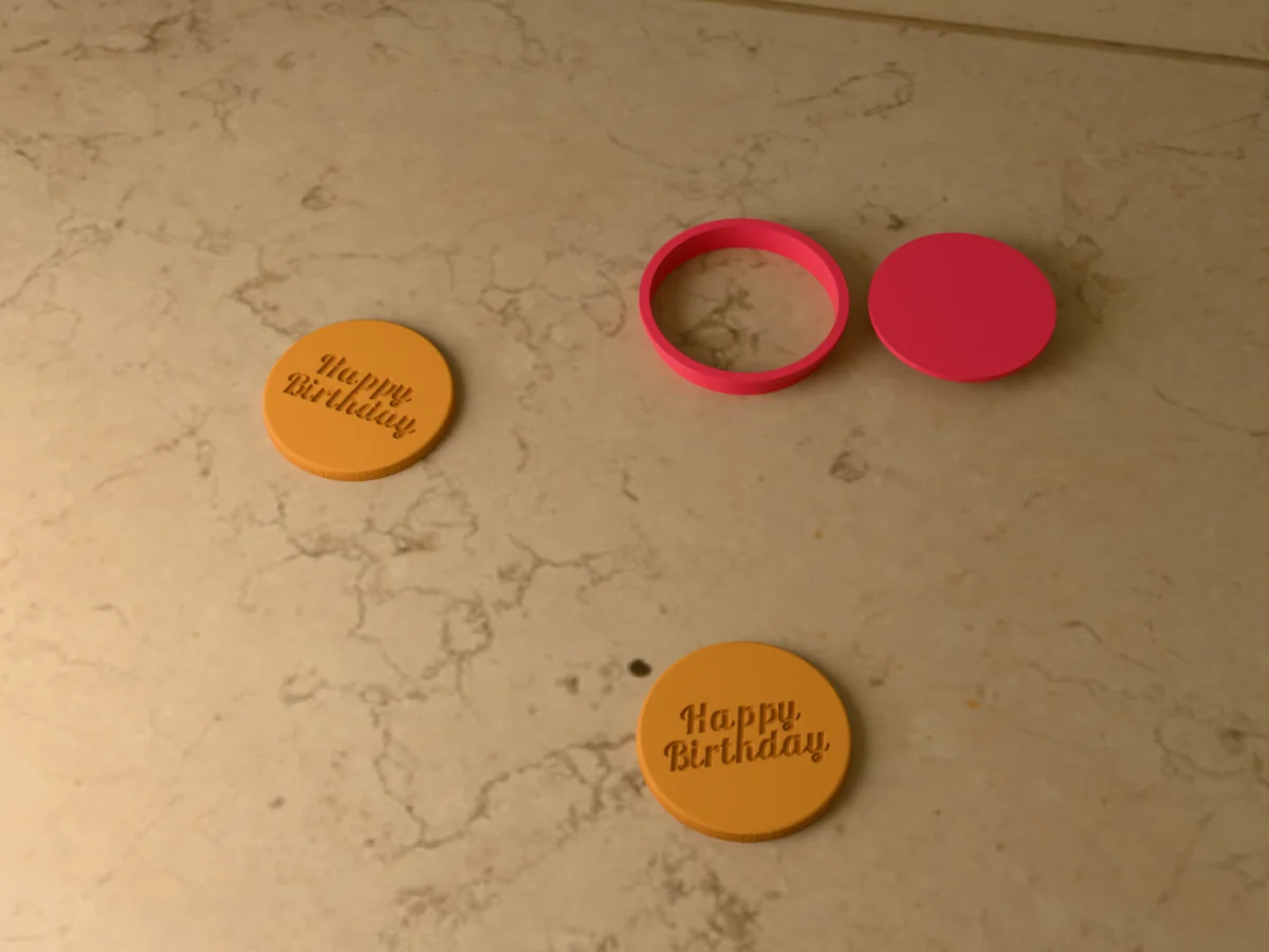 3D render of a pink 3D-printed round cookie cutter and Happy Birthday stamp next to two golden baked cookies with Happy Birthday pressed in
