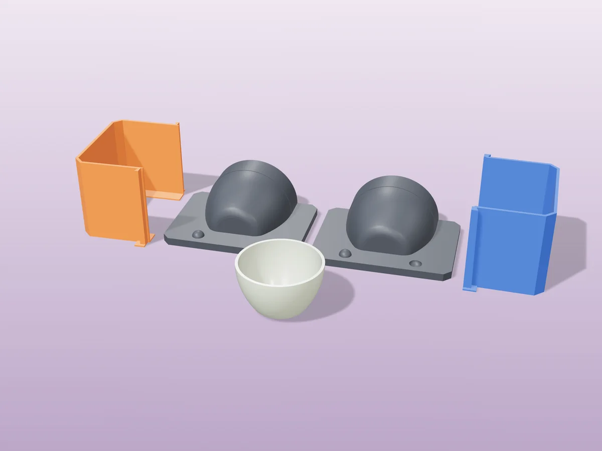 3D render of two printed plaster-mold housings for a matcha bowl, orange and blue shells beside charcoal bases, with a sage green fired tea bowl in front