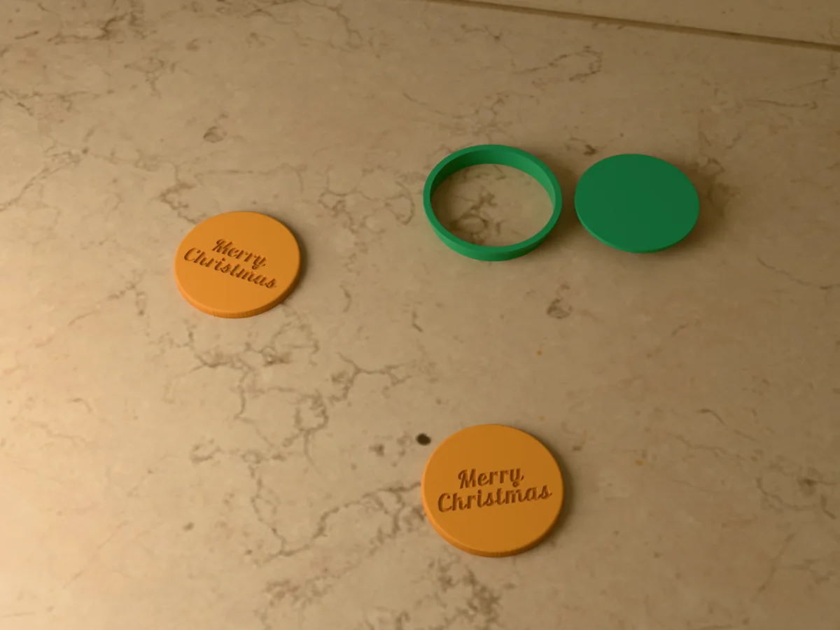3D render of a green 3D-printed round cookie cutter and Merry Christmas stamp next to two golden baked cookies with Merry Christmas pressed in