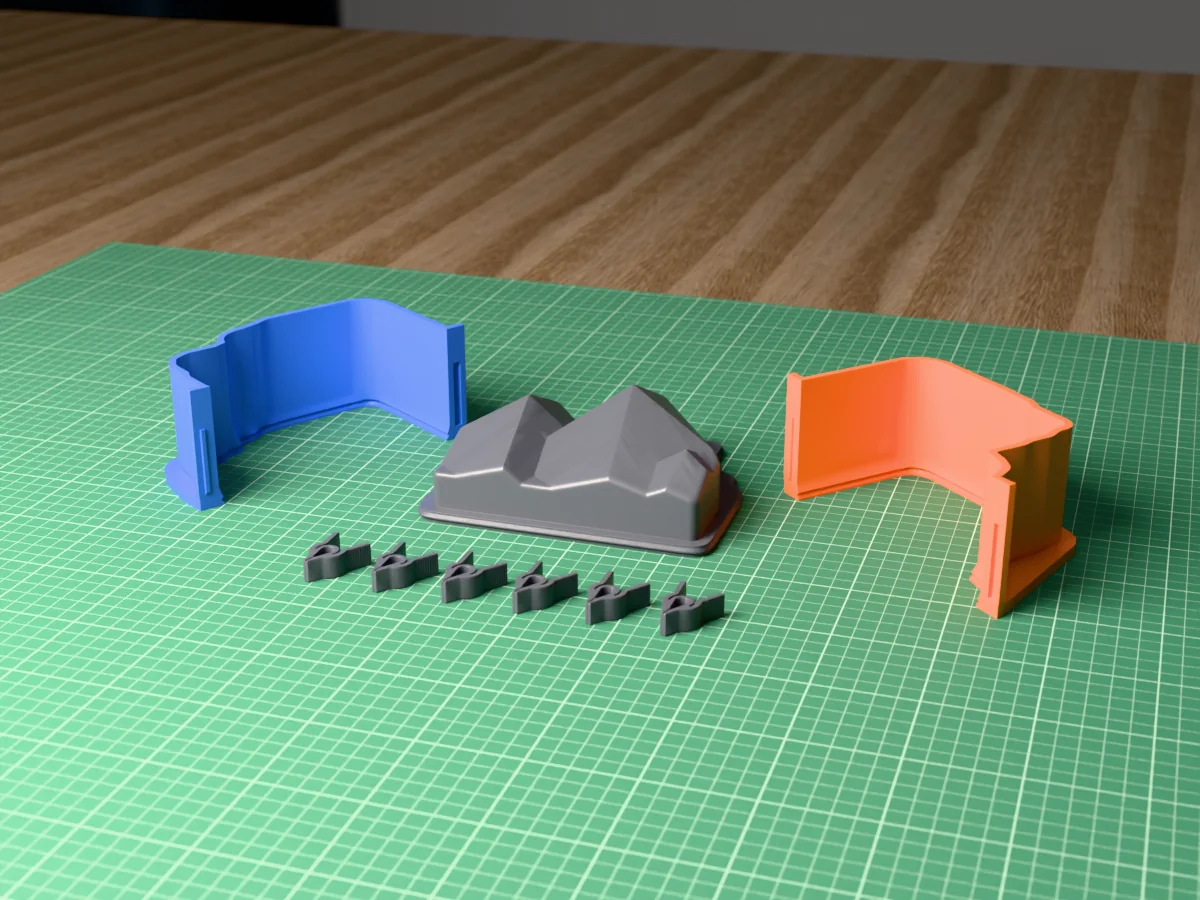 3D render of the faceted low-poly mountain bookend master lying on its back, fused on its printed charcoal base between the orange and blue shell halves, with the charcoal clamps in front