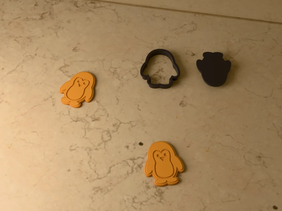 3D render of a black 3D-printed penguin cookie cutter and stamp next to two golden baked cookies with the face, belly, eyes and beak pressed in