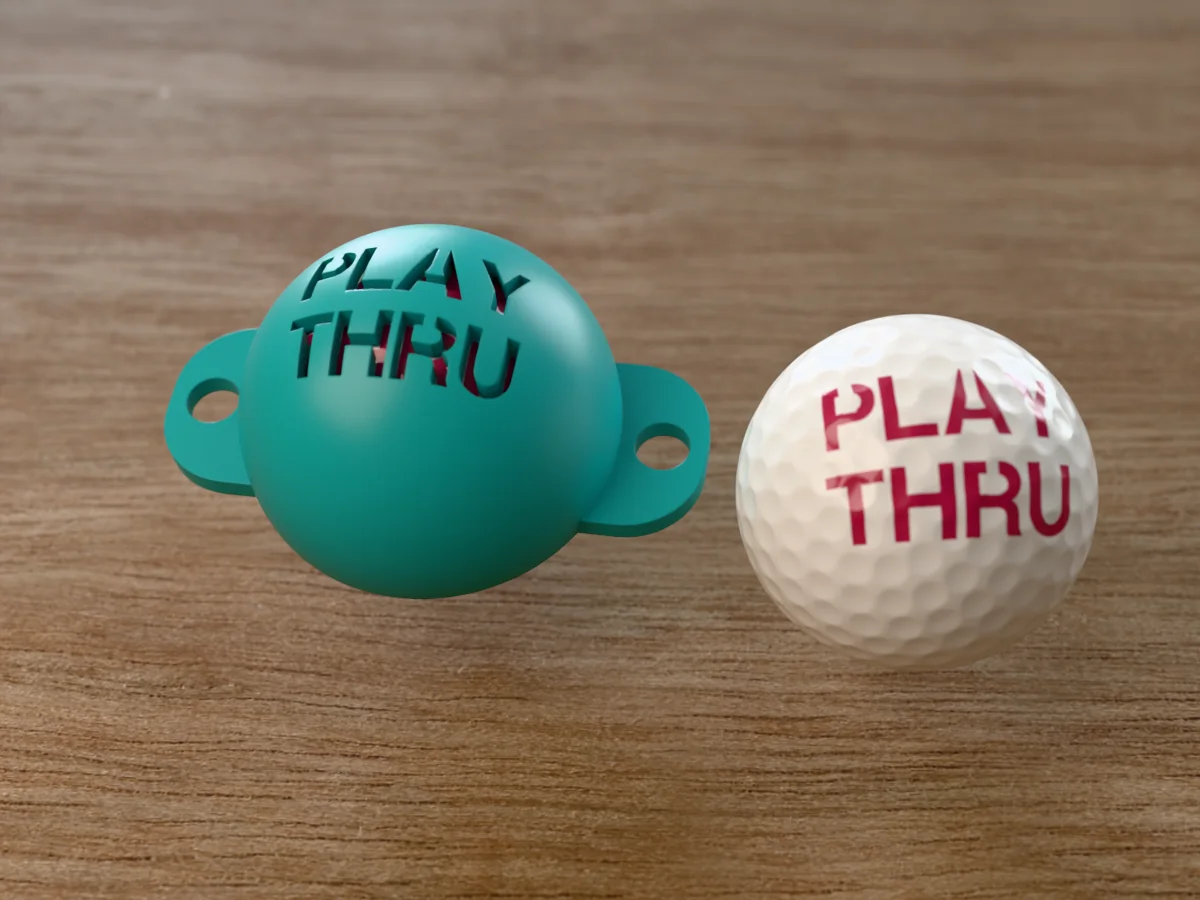 3D render of a teal 3D-printed golf ball stencil with PLAY THRU cut through it, sitting on a golf ball, next to a ball marked with PLAY THRU in pink