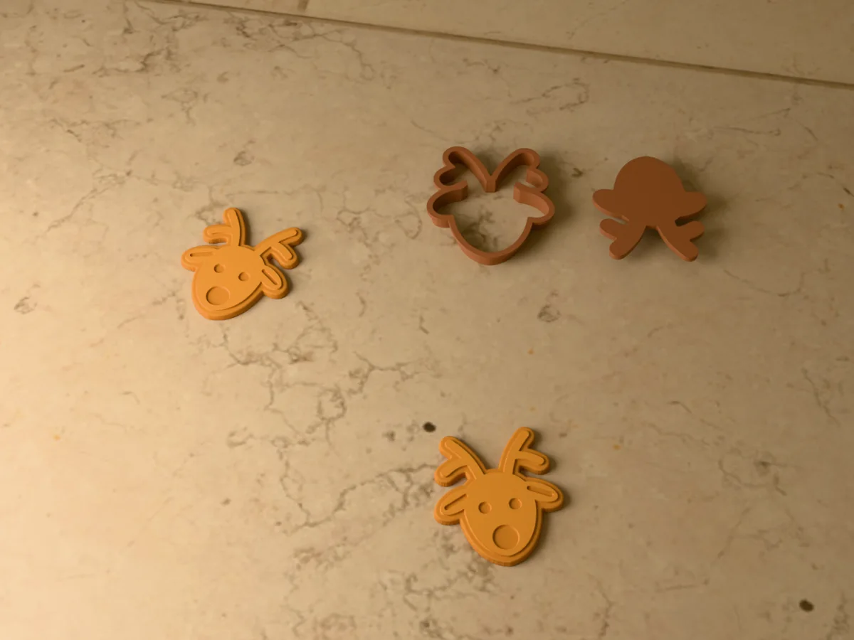 3D render of a brown 3D-printed reindeer cookie cutter and stamp next to two golden baked cookies with the eyes, ears and a round nose pressed in