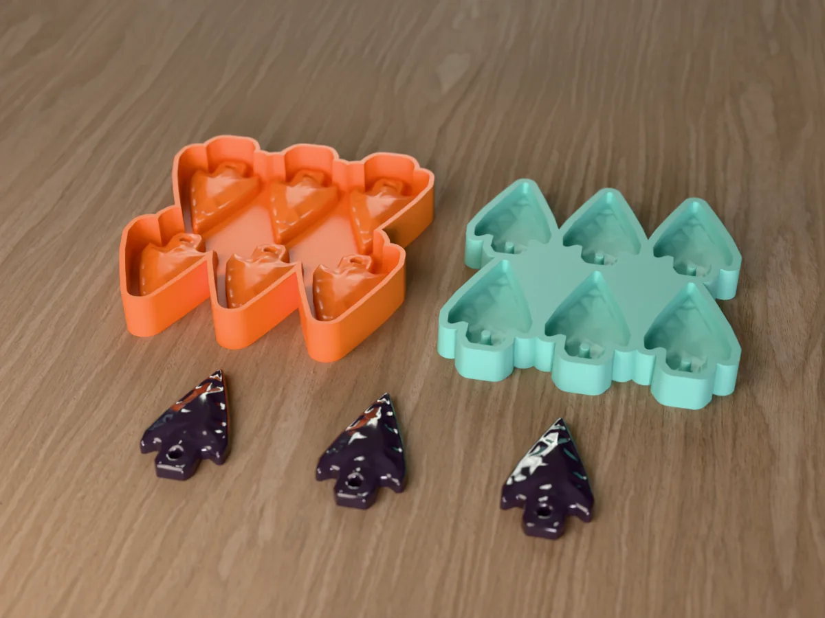 3D render of the printed caster for a six-cavity resin arrowhead pendant mold beside the teal silicone tray it pours, with glossy dark resin arrowhead pendants in front