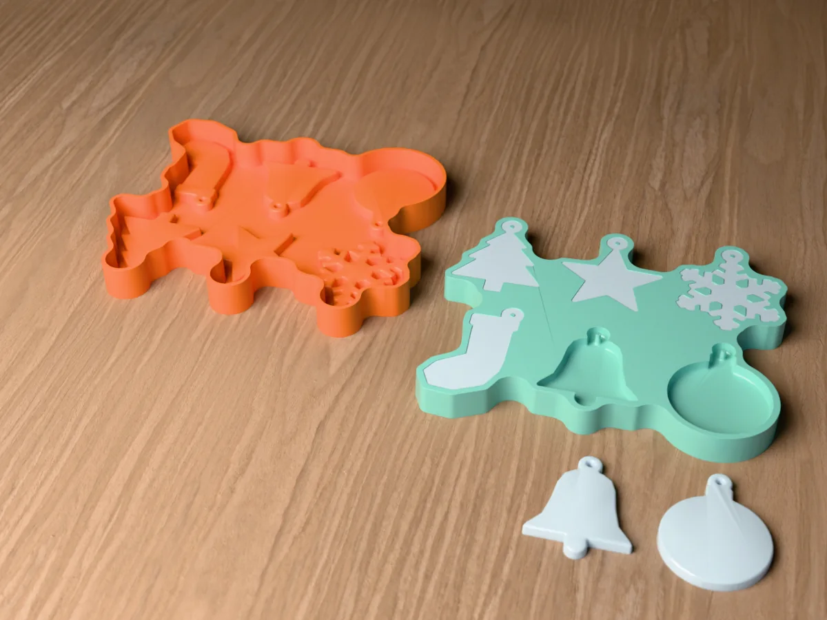 3D render of an orange printed caster beside a green silicone resin Christmas ornament mold holding pale blue tree, star, snowflake and stocking ornaments, with a bell and a bauble ornament in front