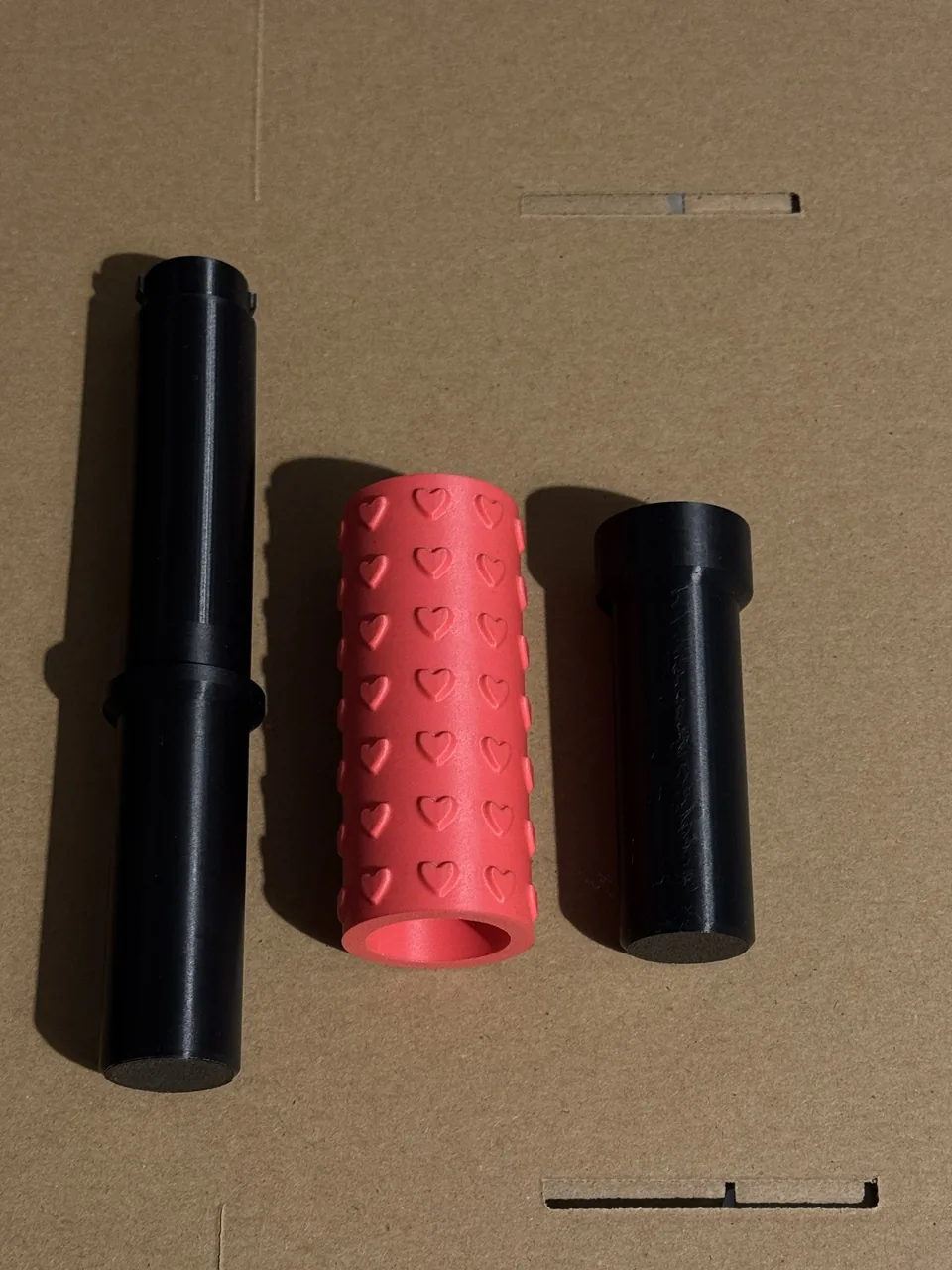 Photo of the three printed parts side by side: the black core handle with its locking lugs, the coral hearts sleeve and the black cap handle