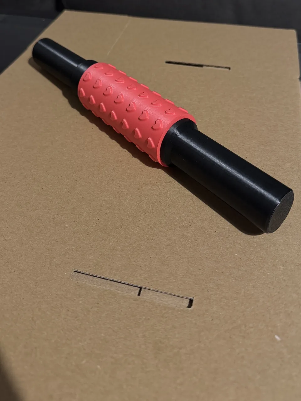 Photo of a 3D printed textured rolling pin: a coral sleeve with raised hearts on two black handles, lying on cardboard
