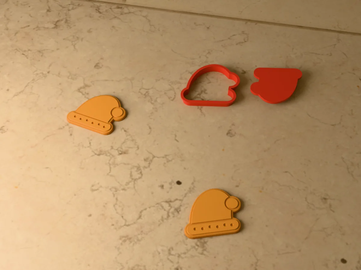 3D render of a red 3D-printed Santa hat cookie cutter and stamp next to two golden baked cookies with the fur brim and the pompom pressed in