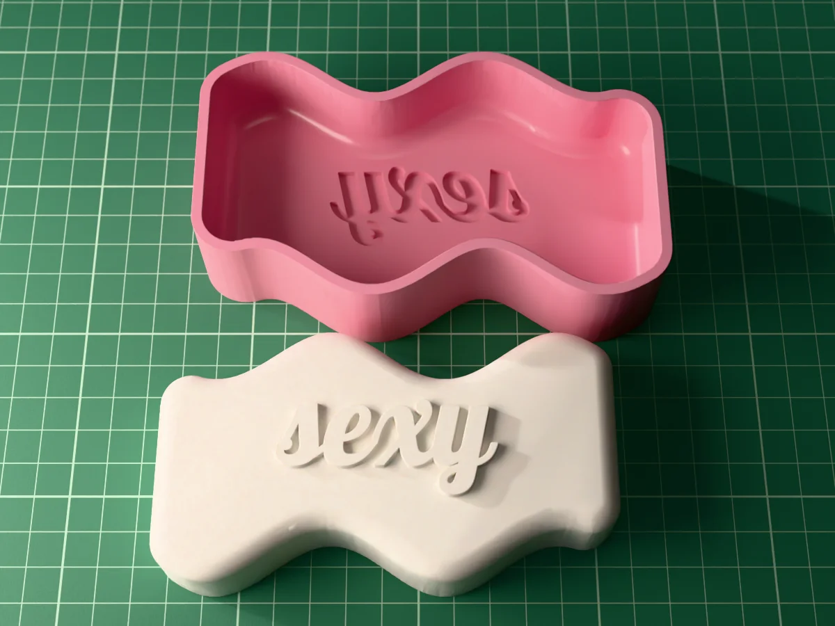 3D render from above of a white wavy slab candle lying face up with "sexy" in raised script, the silicone mold it is cast in behind it