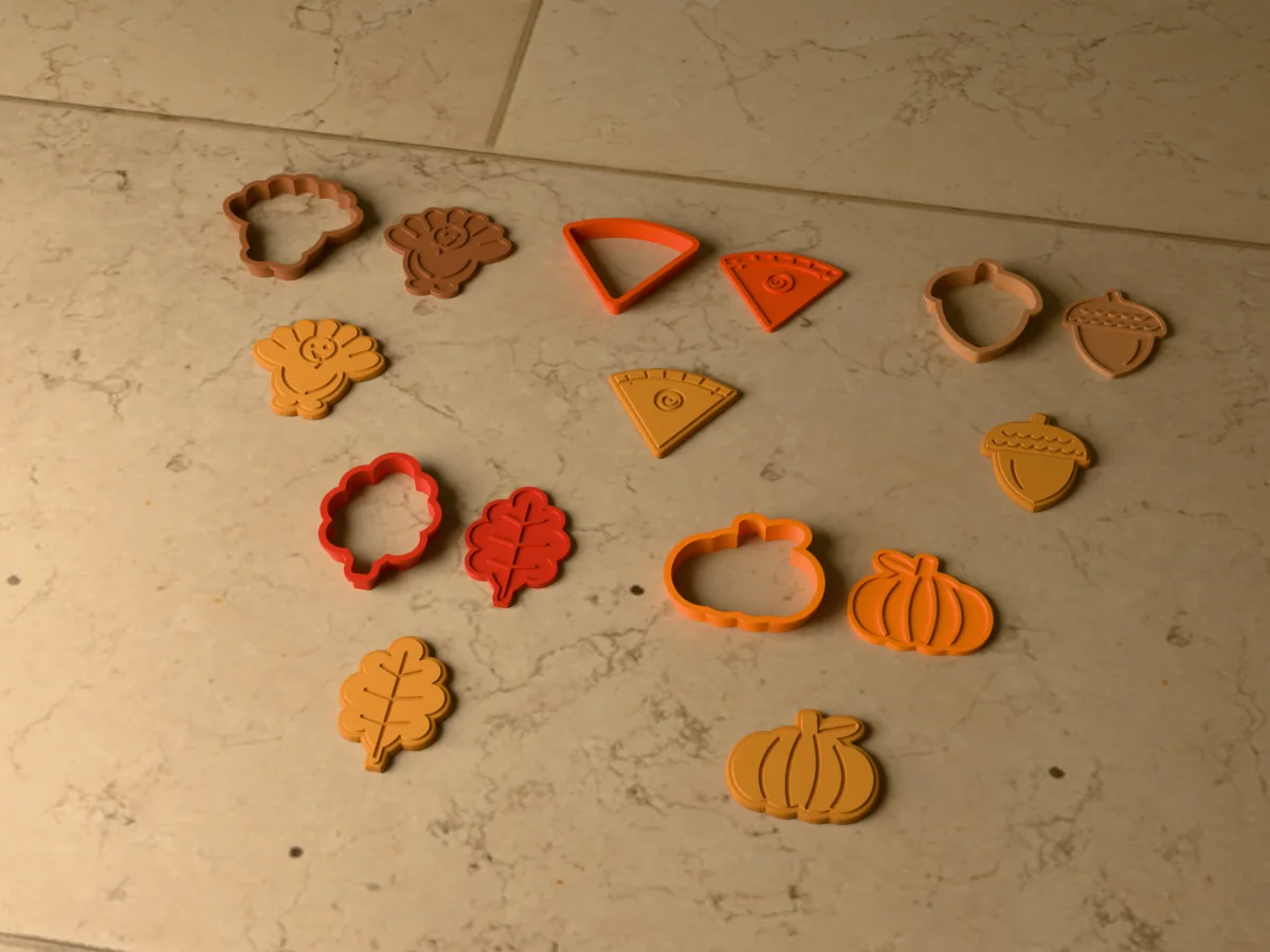 3D render of five 3D-printed Thanksgiving cookie cutters with their stamps and a golden stamped cookie in front of each: turkey, slice of pie, acorn, oak leaf and pumpkin