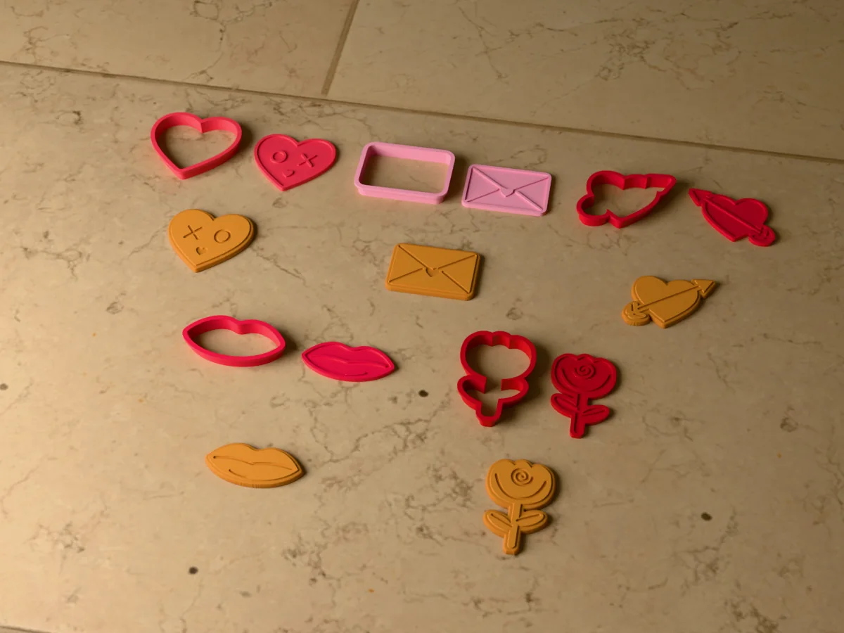 3D render of five 3D-printed Valentine's cookie cutters with their stamps and a golden stamped cookie in front of each: XO heart, love letter, heart and arrow, lips and rose