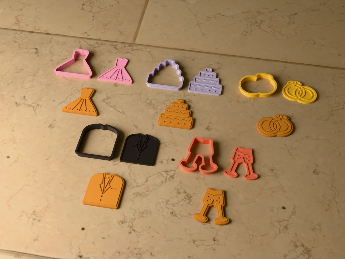 3D render of five 3D-printed wedding cookie cutters with their stamps and a golden stamped cookie in front of each: wedding dress, wedding cake, wedding rings, suit and bow tie and champagne glasses