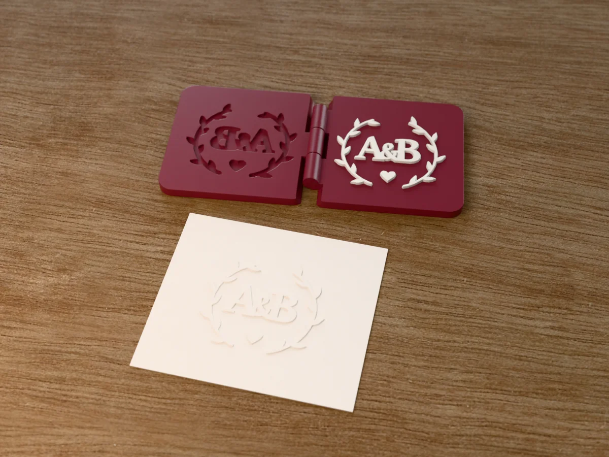 3D render of the printed wedding monogram embosser, open flat on its hinge, with the raised die and the matching cavity, next to a card embossed with its mark