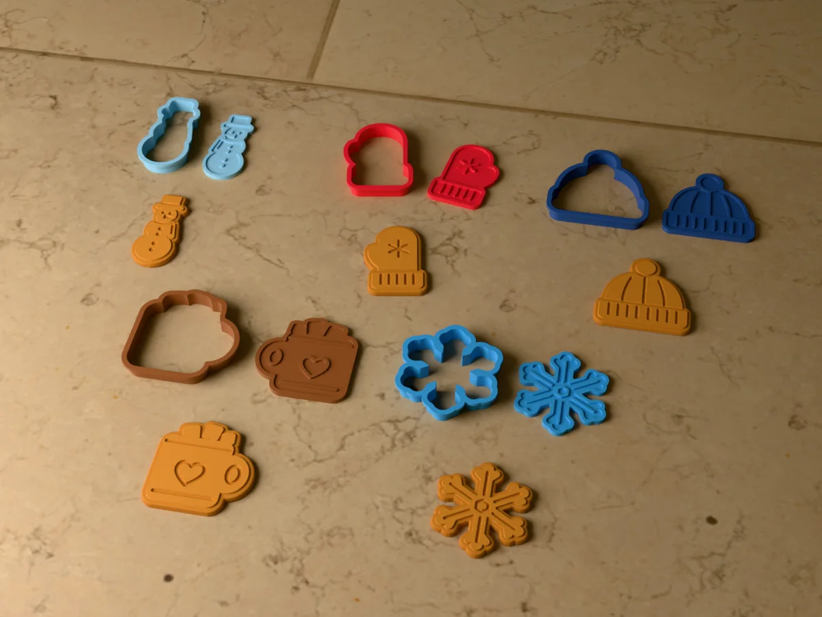 3D render of five 3D-printed winter cookie cutters with their stamps and a golden stamped cookie in front of each: snowman, mitten, beanie hat, mug of cocoa and snowflake