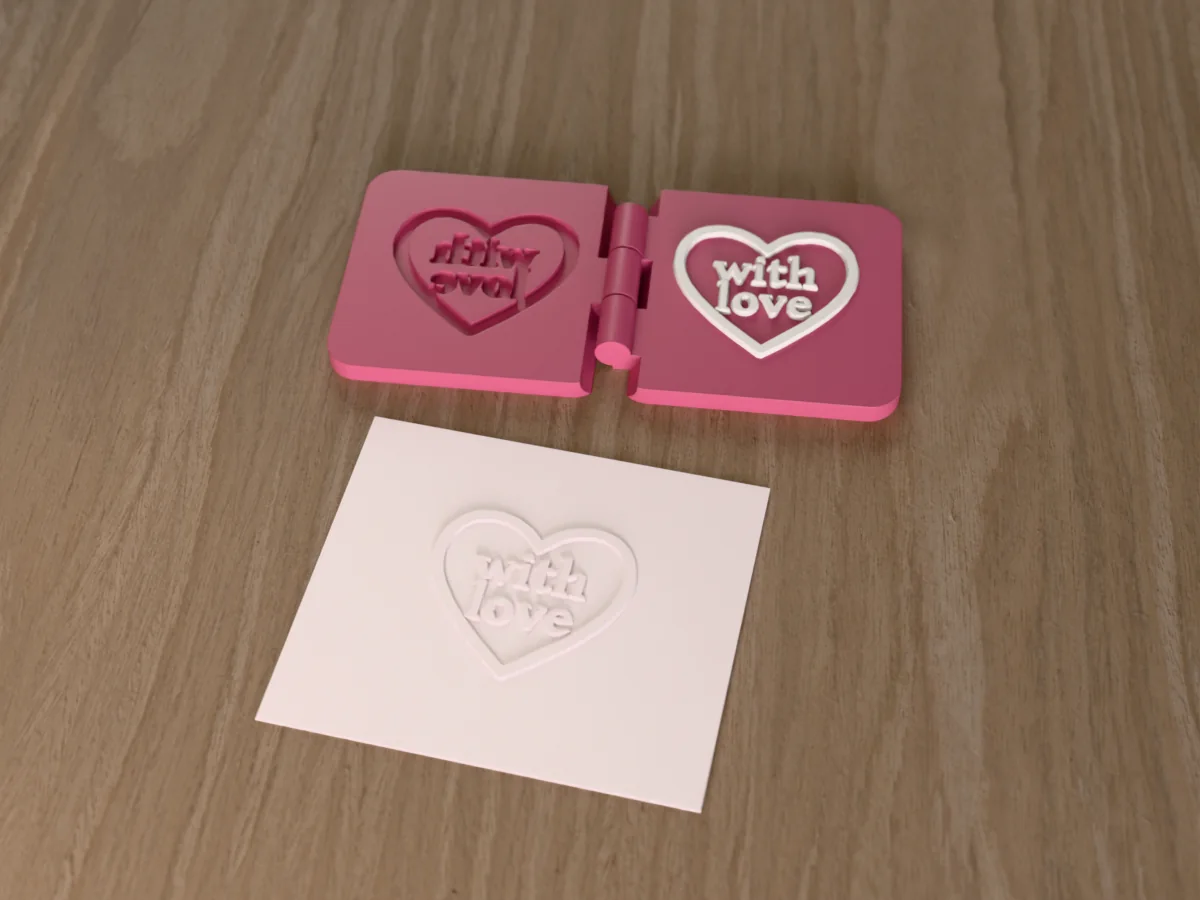 3D render of the printed with love heart embosser, open flat on its hinge, with the raised die and the matching cavity, next to a card embossed with its mark