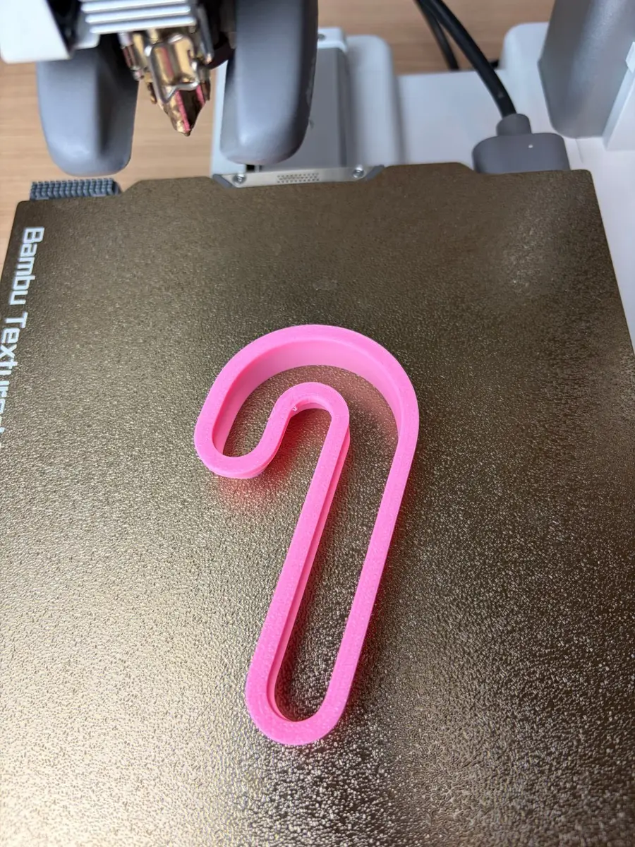 The pink 3D-printed candy cane cookie cutter on a textured print bed, just off the printer