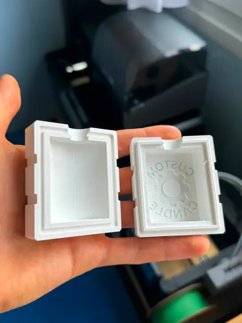 Two printed halves of a small candle mold held open in one hand, the cavity of each clearly visible