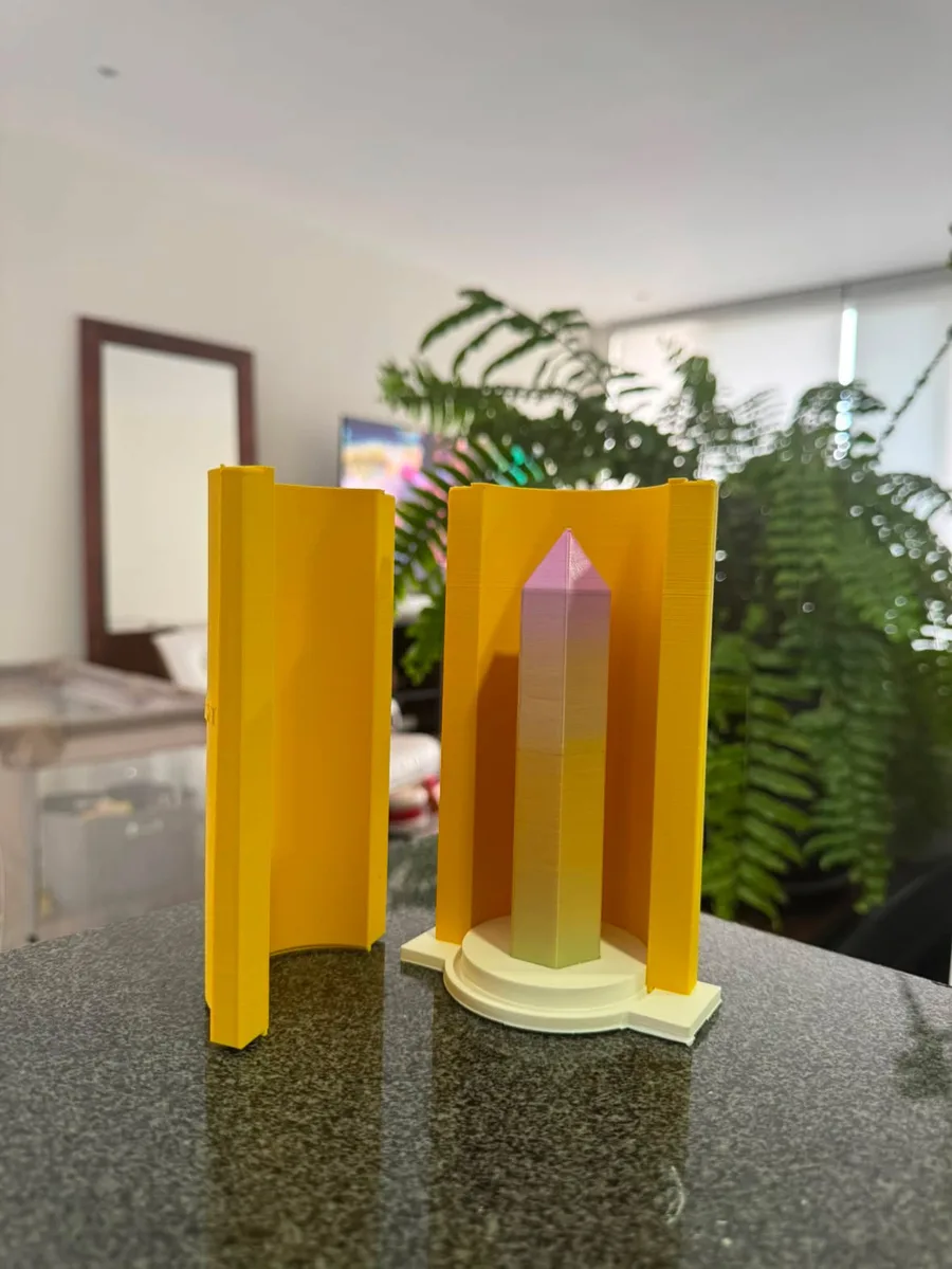 A 3D-printed pink-to-yellow crystal tower standing on its base plate between the two open halves of the gold printed formwork that gets filled with silicone around it