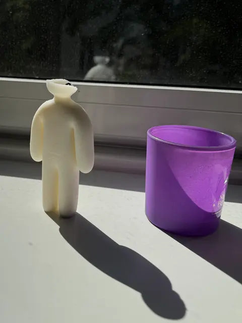 A cream wax figure candle standing in the sun on a windowsill next to a purple candle jar, cast in a 3D-printed two-part mold.