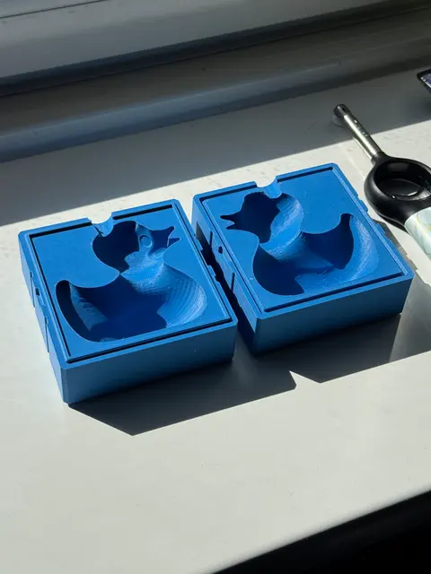 Both halves of a blue 3D-printed two-part rubber-duck mold lying open on a windowsill, each showing its carved cavity and the notch the wax pours through.