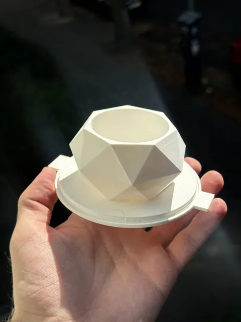 A white 3D-printed faceted plant pot master on its round base plate, held in one hand at a window, ready for the mold walls to seat around it.