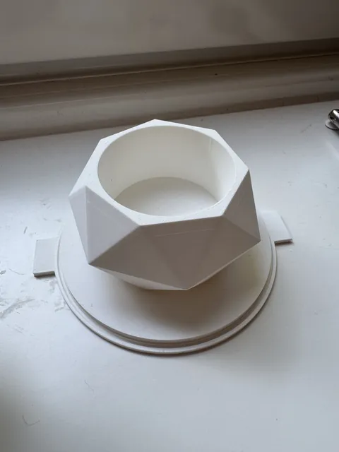 A white 3D-printed faceted plant pot master standing on a windowsill, the seating groove that the mold wall drops into running right around its round base plate.
