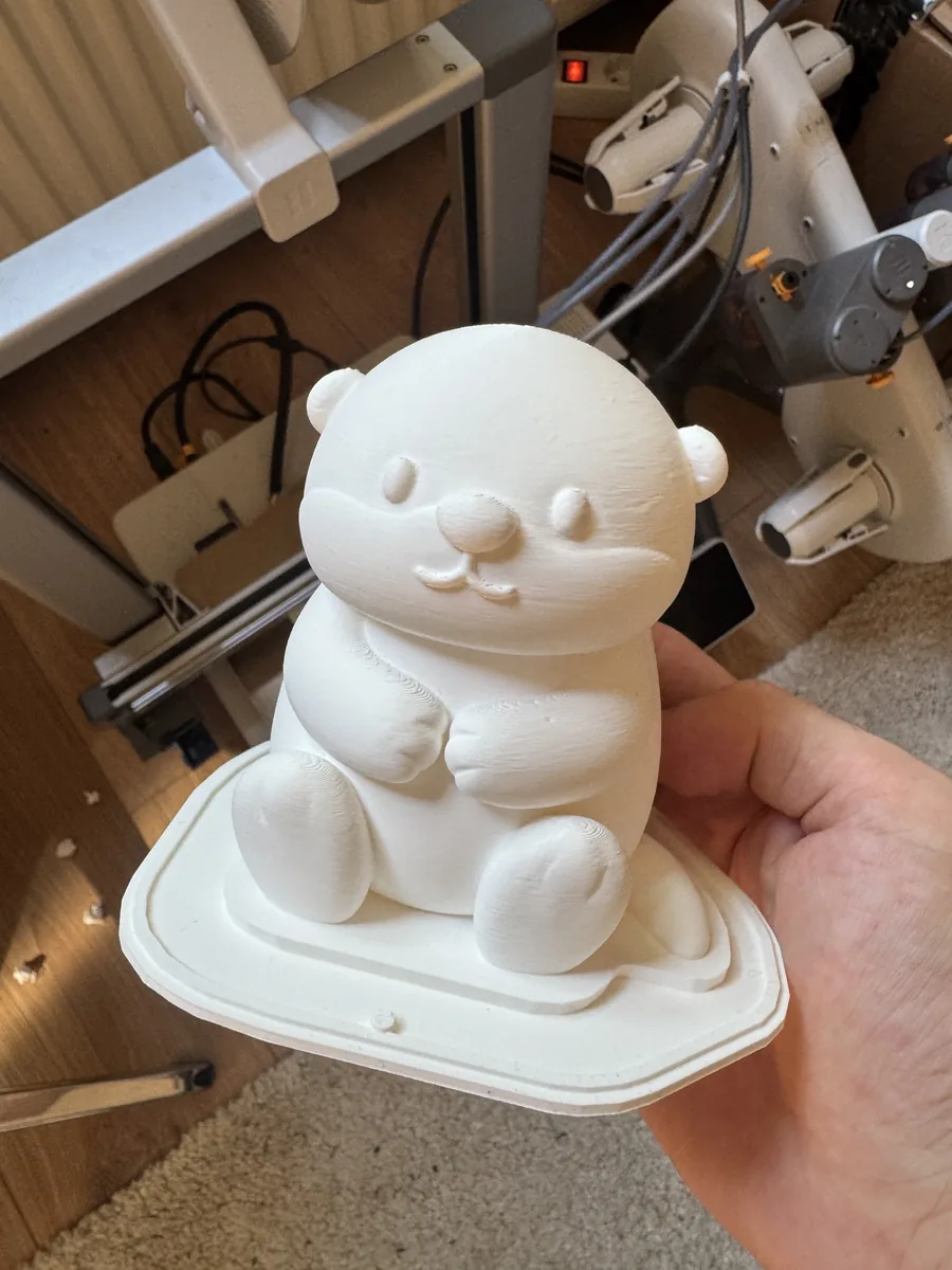 A white 3D-printed sitting bear figure master on its rounded-square mold base plate, held up in front of the 3D printer that produced it