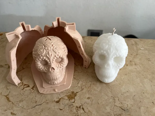 A salmon 3D-printed two-part candle mold open behind its printed sugar-skull master, with the finished white wax skull candle standing beside it.