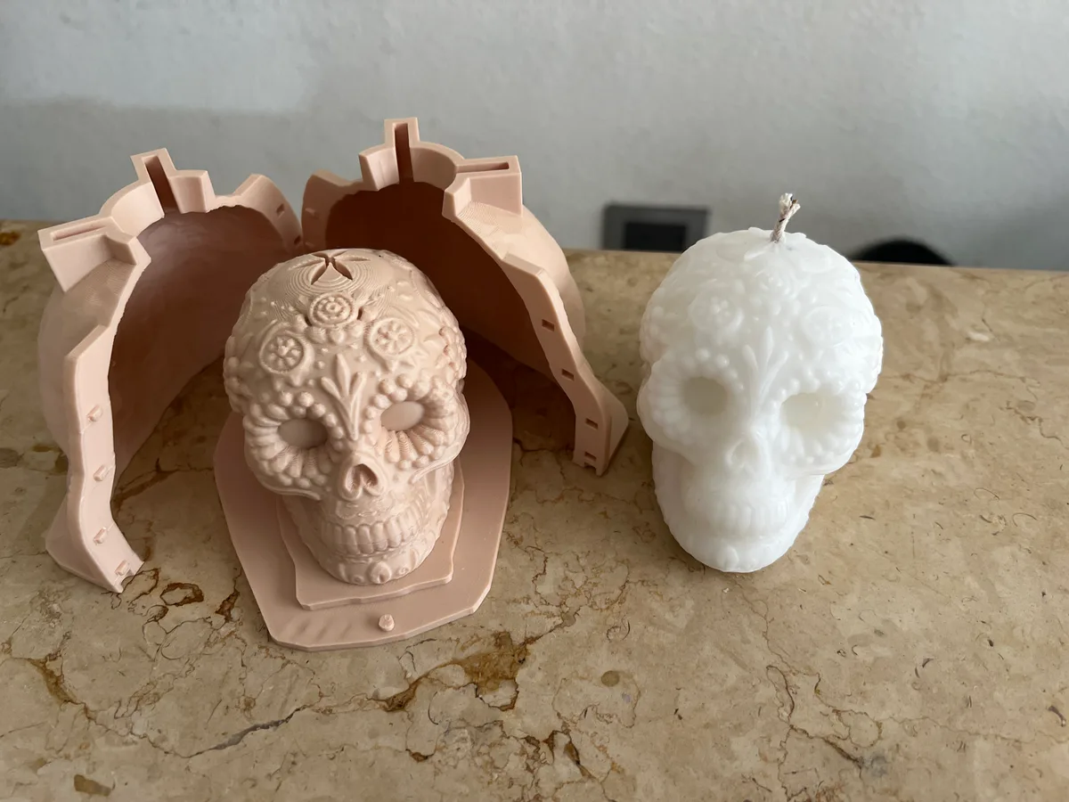 A salmon 3D-printed two-part candle mold standing open around its printed sugar-skull master, with the finished white wax skull candle beside it