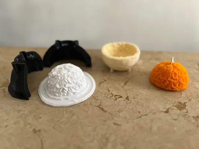 A black 3D-printed two-part mold beside a white marigold-dome master, the pale wax shell it cast, and the finished orange marigold candle.