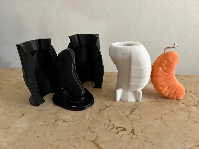 A black 3D-printed two-part mold standing open with its master, the white cast beside it, and the finished orange wax candle.