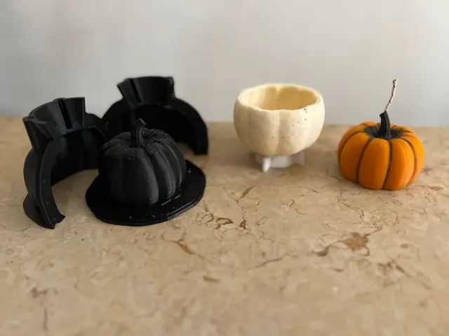 A black 3D-printed two-part pumpkin mold with its printed master, the pale wax shell it casts, and the finished orange pumpkin candle.